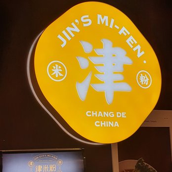 Jin’s Mi-Fen - ows a neon sign for a chinese restaurant - The image shows a bright yellow, illuminated circular sign with the text 'Jin's Mi-Fen' at the top and 'Chang De China' at the bottom. The sign includes Chinese characters and symbols, suggesting a Chinese cuisine restaurant. The sign is mounted indoors, with a dark background, and part of a menu or advertisement is visible below it, along with a vending machine stocked with canned beverages. - Chinese Near Me - Markham, Ontario