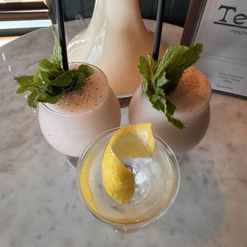 Teddy's - Three drinks are placed on a marble table at Teddy's in Austin, TX. Two are pink cocktails garnished with fresh mint leaves and black straws, while the third is a clear drink with a lemon twist. A decorative lamp and a framed menu are visible in the background, indicating a cozy, dark-ambiance bar setting. - Cocktail Bars Near Me - Austin, Texas