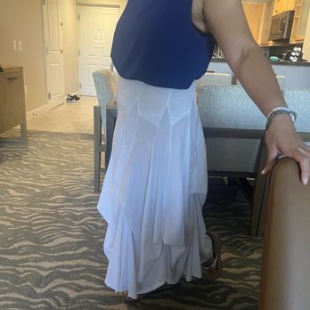 Pretty Please Boutique - Sale top - A woman is standing in a hotel room, wearing a sleeveless blue top and a long, flowing white skirt. Her right arm is resting on a chair, and she is wearing a bracelet. The room features a patterned carpet, a dining table with chairs, and a kitchenette in the background. The door and some shoes are visible near the entrance. - Women's Clothing Near Me - Phoenix, Arizona