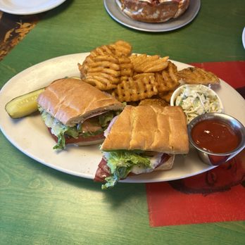 Oregano's - Original Italian stuffed sandwich - A plate contains two sandwiches with lettuce, tomato, and cheese on toasted bread, accompanied by waffle fries, coleslaw, and a pickle. In the background, there is a small pizza with toppings including pepperoni and diced tomatoes on a plate. The setting appears to be a casual dining environment with a green table surface. - Pizza Near Me - Gilbert, Arizona