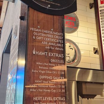 Hawt Pizza Co. - The image shows a tall, wooden menu board inside Hawt Pizza Co., featuring various offerings. The menu includes vegan cheese, gluten-free crust, gift certificates, and extras such as drizzles and dipping sauces. Prices are listed for items like Mike's Hot Honey, balsamic glaze, extra virgin olive oil, and various ranch and honey cup options. Additional items include post-oven arugula and a burrata ball. The setting appears to be inside a modern food hall with tiled walls and a stainless steel counter nearby. - Pizza Near Me - Albuquerque, New Mexico