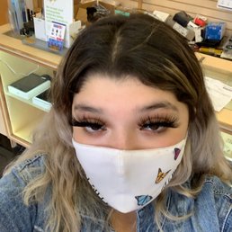 Purrr - A woman with long, wavy hair and false eyelashes wears a butterfly-patterned face mask and denim jacket, taking a selfie inside a retail store.