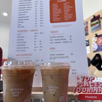 Window Coffee Bar - Americano and latte - Two iced coffee drinks in clear plastic cups with the Window Coffee Bar logo are positioned in front of a menu board. The menu lists various coffee beverages, teas, toasts, and food items, with prices. The drinks include signature lattes, espresso, and non-espresso options. The menu also features flavors, milk options, and food choices like turkey sandwiches and bagels. The setting appears to be inside the coffee shop, with the drinks placed on a glass surface. - Coffee & Tea Near Me - Phoenix, Arizona