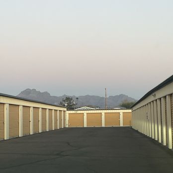 Anytime Storage - The view of our purple mountains majesty (the Superstition Mountains) from the units at Anytime Storage. - The image depicts a row of storage units with beige roll-up doors, situated outdoors on a paved surface. The units are aligned on both sides, creating a corridor leading towards a mountain range visible in the distance. The sky is clear, and the lighting suggests it is either early morning or late evening. The scene is neutral and focused on the storage facility with natural landscape in the background. - Self Storage Near Me - Apache Junction, Arizona