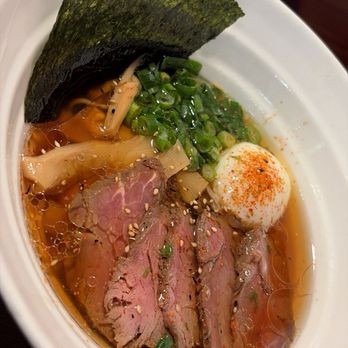 Strings Ramen - Lakeview - Miso Premium - A bowl of ramen contains sliced beef, a soft-boiled egg with a sprinkle of red seasoning, chopped green onions, bamboo shoots, and a sheet of seaweed. The broth appears rich with oil droplets on the surface, and the ingredients are arranged to showcase their colors and textures. - Ramen Near Me - Chicago, Illinois
