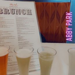 Mimosa flight. This was delicious. The only thing I can fully recommend. - Four mimosas in glasses are arranged on a table at Abby Park restaurant, with a brunch menu visible in the background and a neon sign reading 'Abby Park' on the window.