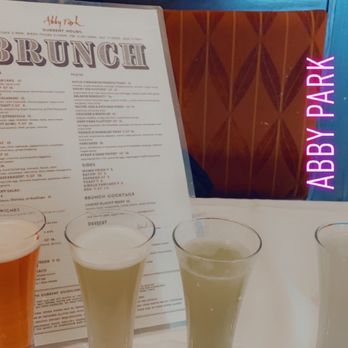 Abby Park - Mimosa flight. This was delicious. The only thing I can fully recommend. - Four mimosas in glasses are arranged on a table at Abby Park restaurant, with a brunch menu visible in the background and a neon sign reading 'Abby Park' on the window. - New American Near Me - Milton, Massachusetts