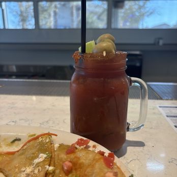 Hash Kitchen - Bloody Mary - A Bloody Mary drink with celery, olives, and a salted rim in a mason jar, placed on a counter near a plate of food. - New American Near Me - Gilbert, Arizona