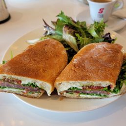 Prosciutto Di Parma Sandwich - A white plate holds two half sandwiches with toasted bread, filled with meats, lettuce, and other vegetables, served alongside a mixed green salad. In the background, there are condiments, a cup of coffee, and another sandwich on a plate, indicating a casual dining setting at a restaurant.