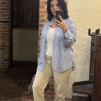 Zinnia - Me - A woman with shoulder-length dark hair wears a striped blue shirt over a white top and beige pants, taking a mirror selfie in a space with brick and textured walls. - Cocktail Bars Near Me - Silver Spring, Maryland