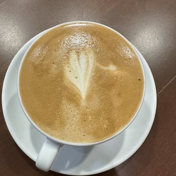 Chess Club Coffee - A coffee drink - A close-up view of a coffee drink with foam art, displaying a leaf pattern on the surface. The cup is white and sits on a matching saucer, placed on a dark wooden table. The foam art is centered and detailed, highlighting the barista's skill in latte art. - Coffee & Tea Near Me - Chicago, Illinois