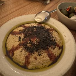 Babaganoush - A bowl of baba ganoush garnished with black seasoning, surrounded by olive oil, on a wooden table with other dishes and utensils.