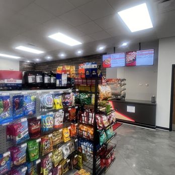 Clay Texaco - Snacks - The image shows snack shelves stocked with a variety of chips and snack foods, positioned near digital menu screens displaying food options. The setting appears to be inside a convenience store or gas station, with a tiled ceiling and bright overhead lighting. The digital screens are mounted on a wall behind the snack display, and the store has a modern interior design with a brick accent wall in the background. - Convenience Stores Near Me - Birmingham, Alabama