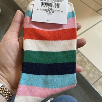 LOFT - A person is holding a single sock with horizontal stripes in multiple colors, including pink, red, white, green, navy, and light blue. The sock has a tag attached with product details, including material composition and care instructions. The background shows a tiled floor and part of the person's lap, indicating they are seated in a retail or shopping environment. - Accessories Near Me - Tampa, Florida