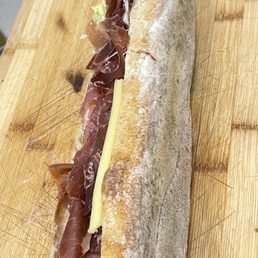 A baguette sandwich with prosciutto, cheese, and lettuce is placed on a wooden cutting board, with a background of a clinical or kitchen setting.