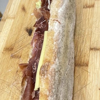 Nouvelle Maison - A baguette sandwich with prosciutto, cheese, and lettuce is placed on a wooden cutting board, with a background of a clinical or kitchen setting. - Grocery Near Me - Winchester, Massachusetts