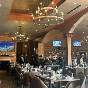 Divan Restaurant - Nice place and see I can watch the game if I wanted! - The image shows a restaurant interior with a warm color palette, including brick walls and a wooden floor. Several tables are arranged with wine glasses, napkins, and cutlery, ready for guests. Large chandeliers with candle-like lights hang from the ceiling, providing ambient lighting. In the background, a bar area displays bottles of alcohol on shelves, with two TV screens mounted on the brick wall. Waitstaff are present, and the overall setting suggests a formal dining environment. - Persian/Iranian Near Me - Mclean, Virginia
