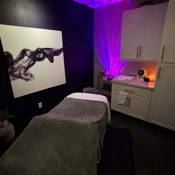 Fuchsia - Ahwatukee - A massage room features a massage table with a gray cover, abstract black and white wall art, and purple lighting effects projected onto the ceiling and corner. - Day Spas Near Me - Phoenix, Arizona
