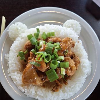 Tasty Pot - Child chicken plate. - A white plate contains a serving of steamed white rice topped with fried chicken pieces coated in sauce. The dish is garnished with chopped green onions. The plate is placed on a dark wooden table, with some paper bags visible in the background. - Hot Pot Near Me - Albuquerque, New Mexico
