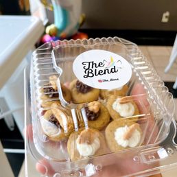 Donuts - A clear plastic container displays mini donuts topped with cream and chocolate, labeled with The Blend's logo and Instagram handle. The background shows part of the café interior with colorful decorations and a counter, indicating a casual dining environment.