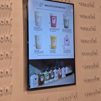 Machi Machi - The digital menu screen at Machi Machi in Markham, ON, features a list of Machi's favorite drinks, including milk teas, smoothies, and a strawberry drink. The display shows images of each beverage with their names and descriptions, and a row of bottled drinks is visible at the bottom of the screen. The background wall has the 'machi' logo repeatedly embossed. - Bubble Tea Near Me - Markham, Ontario