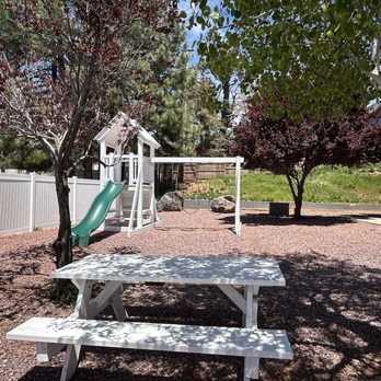 Windmill Coffee - A backyard features several trees providing shade, a green slide attached to a play structure, a swing set, and a white picnic table with benches. The ground is covered with reddish gravel, and a white fence encloses the yard. In the background, there is a grassy area and part of a house with stairs leading to an upper level. - Coffee & Tea Near Me - Strawberry, Arizona