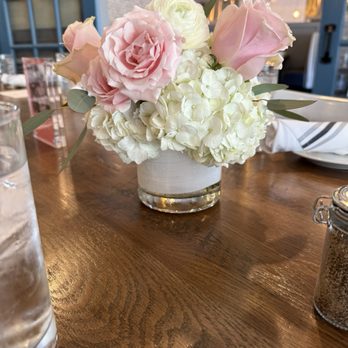 Danielson Flowers - Table Centerpiece - A bouquet of pink roses, white hydrangeas, and greenery in a white vase is placed on a wooden table in a restaurant setting. - Florists Near Me - Shrewsbury, Massachusetts