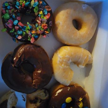 Kim’s Donuts - This establish is more for not so finicky kids.who don't know what a real donut taste like!  Also the chocolate taste weird! - The image shows a white box filled with six donuts, each with distinct toppings and flavors. Some donuts are glazed with vanilla or chocolate, one is decorated with colorful cereal pieces, and another has chocolate icing with M&M candies. The donuts are arranged in two rows, with a partially eaten donut visible at the bottom left. The box is placed on a surface with a piece of paper or napkin nearby. - Donuts Near Me - Lowell, Massachusetts