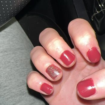 Luxury Nails and Spa - A close-up image of a person's left hand showing manicured nails with red polish and a single glitter accent nail on the ring finger. The hand is positioned inside a vehicle, with part of the car's interior visible, including a black dashboard and a perforated black seat. The person is wearing gray clothing and a blue lanyard with white and orange text. - Hair Removal Near Me - Albuquerque, New Mexico