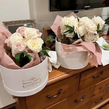 Amazing Flowers - Bridesmaids bouquets - Two flower bouquets with roses and greenery are arranged in white round boxes on a wooden dresser, with a TV and framed photo in the background. - Florists Near Me - Phoenix, Arizona
