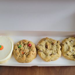 Frosted Sugar Cookie - The image shows four cookies arranged in a pink box. The first cookie is a frosted sugar cookie decorated with a small pumpkin-shaped candy. The remaining three cookies are chocolate chip cookies, each with visible chocolate chunks and a slightly uneven surface. The box is open, revealing the cookies inside, placed on a wooden surface.