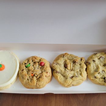 Crumbl - Delaware - Frosted sugar cookie with pumpkin candy, monster, and two milk chocolate chip cookies - The image shows four cookies arranged in a pink box. The first cookie is a frosted sugar cookie decorated with a small pumpkin-shaped candy. The remaining three cookies are chocolate chip cookies, each with visible chocolate chunks and a slightly uneven surface. The box is open, revealing the cookies inside, placed on a wooden surface. - Bakeries Near Me - Delaware, Ohio