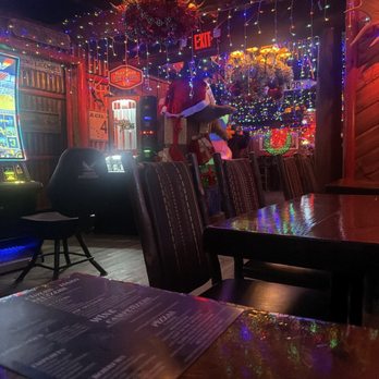 Camp Bar Third Ward - Entry - The image shows the interior of a bar decorated for the holiday season, with colorful string lights, ornaments, and a Santa figure. The ceiling is adorned with multicolored lights and a decorative chandelier. There are tables and chairs, with a menu visible on one table in the foreground. The atmosphere is festive, with holiday-themed decor and lighting creating a warm ambiance. - Bars Near Me - Milwaukee, Wisconsin