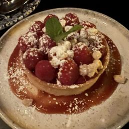 Chocolate tart - A dessert tart featuring a pastry crust filled with fruit, topped with strawberries, raspberries, crumbles, and a mint leaf. The tart is presented on a ceramic plate with a sauce or glaze around it, garnished with powdered sugar or similar topping. The setting appears to be in a dimly lit environment, possibly a restaurant.