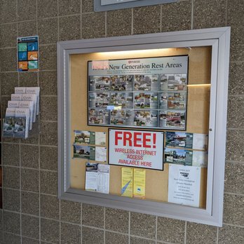 Dows Rest Area - Free! - The image shows a bulletin board mounted on a tiled wall, containing maps, informational notices, and advertisements. A prominent sign in the center announces free wireless internet access available at the location. The board also includes details about Iowa's new generation rest areas, with various smaller notices and brochures around it. A metal plaque above indicates the rest area grounds are maintained by T & L Lawcare. - Rest Stops Near Me - Dows, Iowa