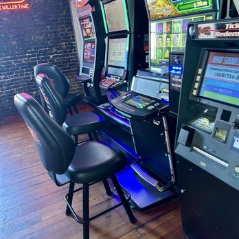 Brewe-Ha's - Games - Multiple slot machines with chairs are arranged against a brick wall in a bar or casino environment, illuminated by colorful lights. - Bars Near Me - Normal, Illinois