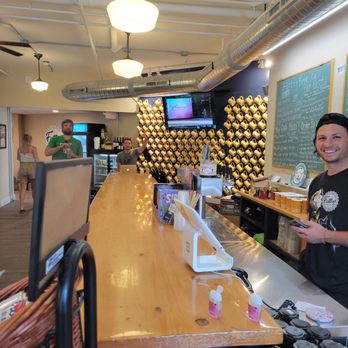 Fattey Beer - The image depicts the interior of Fattey Beer in Buffalo, NY, featuring a long wooden bar counter with staff members behind it. One staff member is smiling and holding a phone, while two other staff members are visible further back, one of whom is waving. The background includes a wall decorated with numerous beer bottles, a mounted TV screen, and a chalkboard menu. The ceiling has exposed pipes and lighting fixtures, creating a casual, industrial ambiance. Patrons are seen walking in the background, contributing to the lively atmosphere. - Beer Bar Near Me - Buffalo, New York