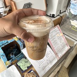Toffee caramel crunch coldbrew with cinnamon - A hand is holding a plastic cup of iced coffee topped with cinnamon at Blissful Brew Coffee Co. The background shows coffee brewing equipment, a menu, and various supplies on the counter, indicating a coffee shop setting. The cup has handwritten notes, and the environment appears cozy and functional for coffee service.