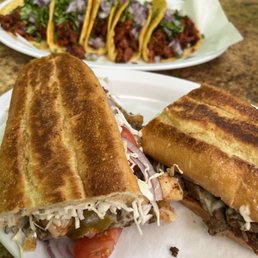 Al Pastor Tacos - Two sandwiches with toasted bread, containing vegetables, cheese, and meat, are placed on a white plate on a brown countertop. In the background, there is a white oval plate with five tacos filled with meat, chopped onions, cilantro, and yellow corn tortillas. The setting appears to be a casual dining environment.