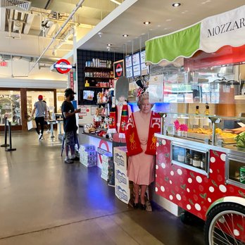 Above Ground - A woman stands behind a food stall decorated with Liverpool FC scarves and merchandise, inside a shopping mall or food court area. The stall has a display case with food items and a sign with the word 'MOZZARELLA'. There are digital menu screens above the stall, and a person is seen browsing nearby. The environment features a high ceiling with exposed pipes and lighting fixtures, and other shoppers are visible in the background. - Fish & Chips Near Me - Washington, DC, District of Columbia