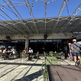 Canopy - The image shows an outdoor seating area of a cafe with a metal roof and string lights. Several tables and green chairs are occupied by people dining, while others stand in line near the entrance. The cafe has large glass windows with signage indicating lunch, dinner, coffee, and takeout options. The sky is clear and blue, and shadows from the roof and string lights are cast on the ground. - Cultural Center Near Me - Austin, Texas