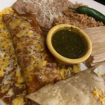 Serranos - Sample plate - A plate of Mexican cuisine includes cheese-covered enchiladas topped with sauce, a serving of rice, refried beans, and a small bowl of green salsa. The dish is served on a white plate with a green chili pepper and a folded tortilla nearby, with shredded cheese sprinkled on some items. - Mexican Near Me - Austin, Texas