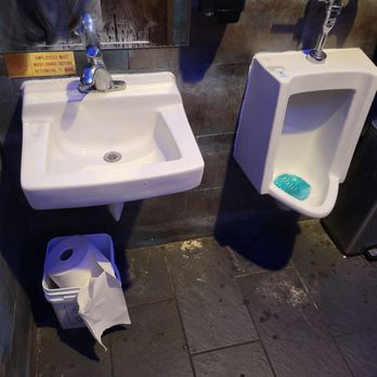 Fancy sushi - The image shows a restroom with a wall-mounted sink on the left and a urinal on the right, both attached to a tiled wall. The sink has a faucet and a mirror above it, with a sign indicating handwashing instructions. Below the sink, there is a trash bin with a paper towel partially hanging out. The urinal has a splash guard at the bottom and a faucet on top. The floor is tiled with dark, textured tiles, and the wall tiles are a mix of dark brown and gray tones. - Sushi Bars Near Me - St. Augustine, Florida