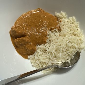 Spice House - A white bowl contains cooked white rice and a serving of curry with a brownish sauce, accompanied by a metal spoon. - Indian Near Me - Edison, New Jersey