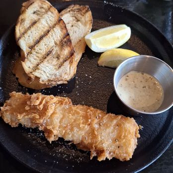 Porter Cullens - A black plate holds a piece of fried fish, two slices of grilled bread, two lemon wedges, and a small metal cup of dipping sauce. The setting appears to be a restaurant table with a glass of water nearby. - Irish Pub Near Me - Evergreen Park, Illinois
