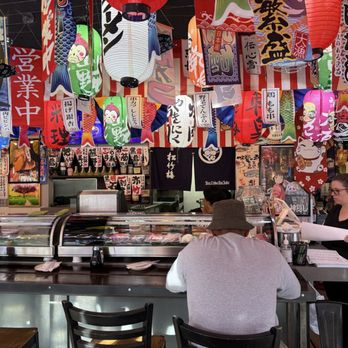 Dragon Sushi - Perfect and joyful decor - The image shows the interior of a Japanese restaurant decorated with numerous colorful paper lanterns and banners hanging from the ceiling. Customers are seated at the counter, with one person wearing a hat and another with their back to the camera. A staff member is standing on the right side, holding a menu or order pad. The background includes Japanese-themed posters and decorations, creating an authentic atmosphere. - Sushi Bars Near Me - Albuquerque, New Mexico