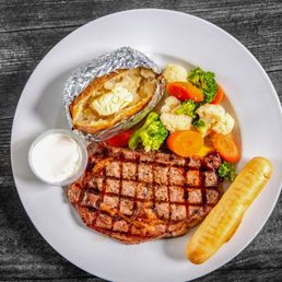 Ribeye Steak - A meal featuring a grilled steak with crosshatch grill marks, a baked potato with butter in foil, mixed steamed vegetables, and a small cup of sour cream.