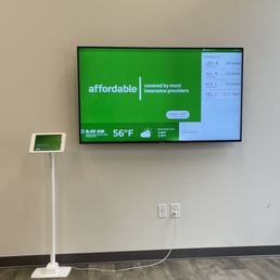 A large flat-screen digital display mounted on a wall shows weather information, current temperature of 56°F, and appointment check-in details for multiple individuals. A smaller tablet device on a stand is positioned to the left of the screen, connected by a cable. The setting appears to be a waiting area or reception in a healthcare or medical office, with a neutral wall and floor visible.