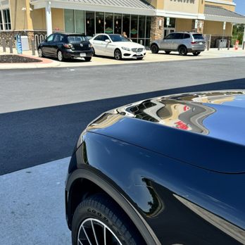 Wawa - The image shows the front part of a black car parked in a Wawa convenience store parking lot. The car's hood reflects the store's sign and part of the building. In the background, there are three other parked cars and the storefront of Wawa, which features large windows and a beige exterior with stone accents. The sky is clear and blue, indicating a sunny day. - Gas Stations Near Me - Sterling, Virginia