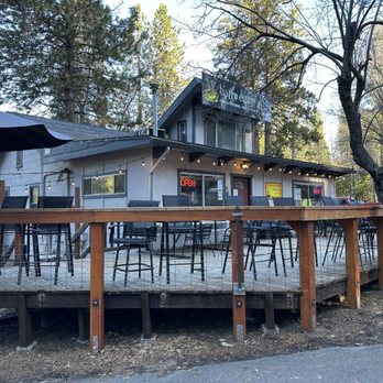 Bistro Espresso - The photo depicts the outdoor patio of Bistro Espresso, with several high tables and chairs arranged on a wooden deck. The deck is elevated and enclosed with a wooden railing. A large umbrella is partially visible on the left side. The building behind the patio has a sign indicating the business name, and there are illuminated signs including an 'Open' sign. The area is surrounded by tall trees, suggesting a location in a wooded or suburban area. The scene is set during daylight with clear weather. - Coffee & Tea Near Me - Arnold, California